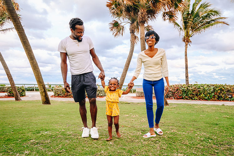  A mother, father, and toddler walking in a field on the shore with palm trees all around.