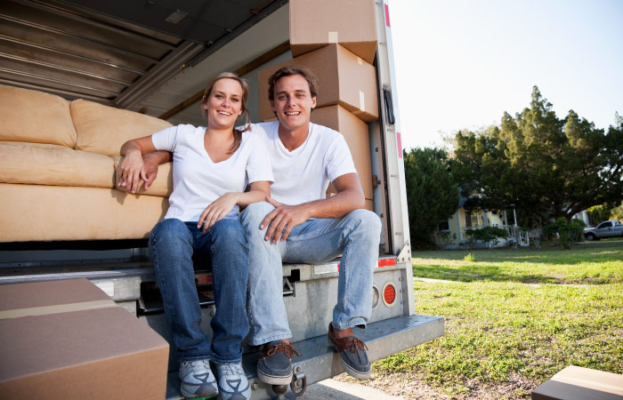 A happy couple is sitting on the back of their moving truck rental. The truck is already loaded with a couch and several moving boxes. 