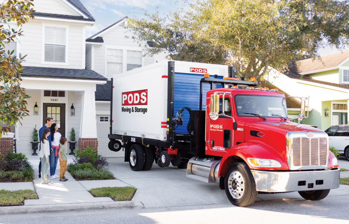 A PODS truck with a moving container on the back is reversed into a residential driveway, as the driver prepares to position the container for easy packing and loading