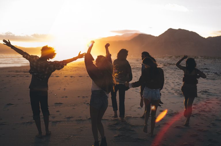 A group of six friends dancing on a California beach during sunset. There’s a mountain range in the distance, and the sun is so bright that the people mostly look like silhouettes.