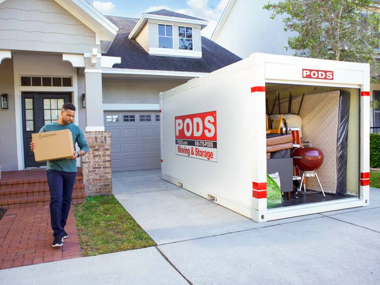 A man loads a box labeled “bedroom” into the PODS container in his driveway.