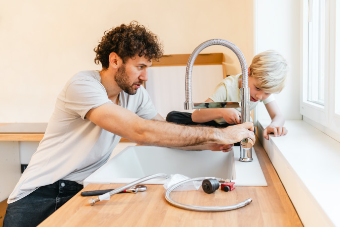 A man is installing a new kitchen faucet as his young son watches