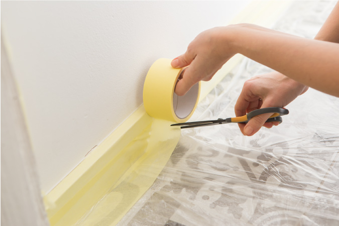 A close up of someone taping off a floor and wall to prep for painting during a DIY kitchen remodel — one hand is holding a roll of painter’s tape while the other hand is cutting it