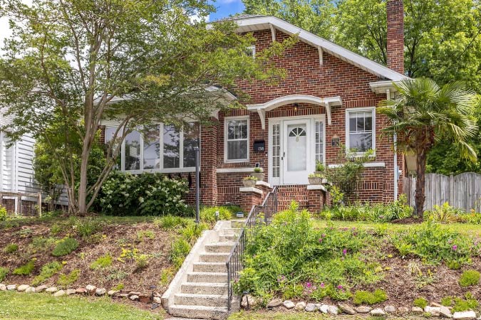 A 1920s brick bungalow on a raised lot in Trinity Park — one of the best Durham neighborhoods — featuring a charming awning above the entrance and concrete steps leading down to street level.
