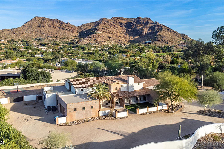 a sprawling mansion in Paradise Valley Village. The left side of the home looks like it’s under construction. There are green trees and mountains in the background. 