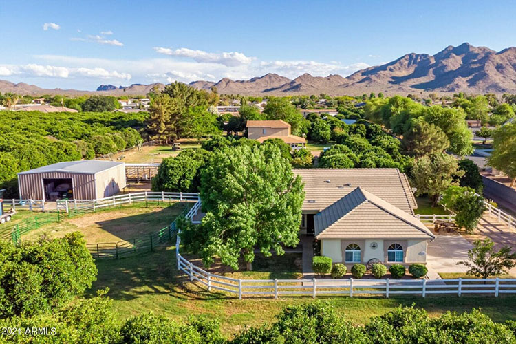 A two-story brown and white home in Queen Creek sits on a few acres of green land with a white fence, with trees and mountains in the background. 