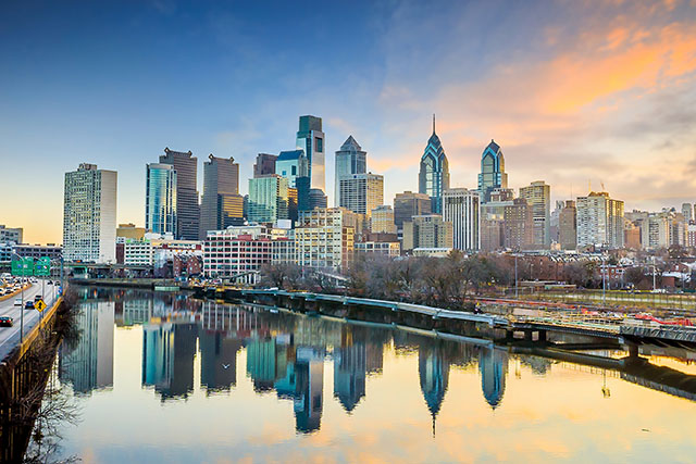 The skyline of Philadelphia, Pennsylvania, as the sun rises. The skyline of high-rise buildings reflects off the water of a river. 
