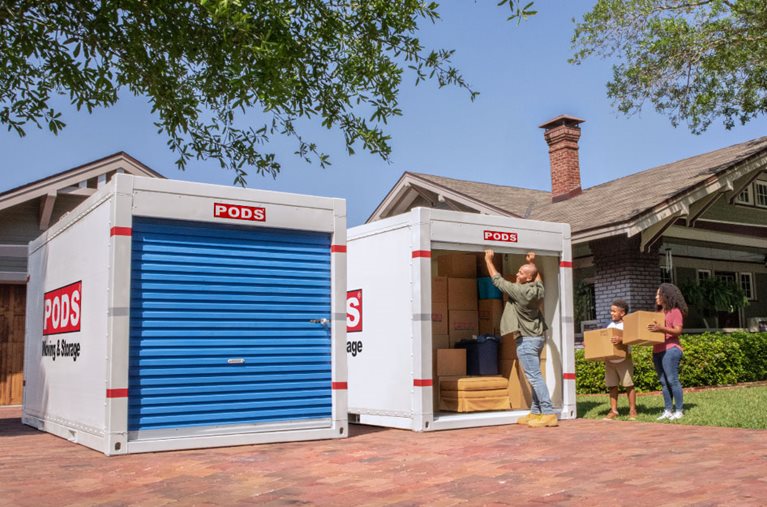A man is opening the door to one of two PODS containers in the driveway as his wife and young son stand by with moving boxes, ready to load into the container.