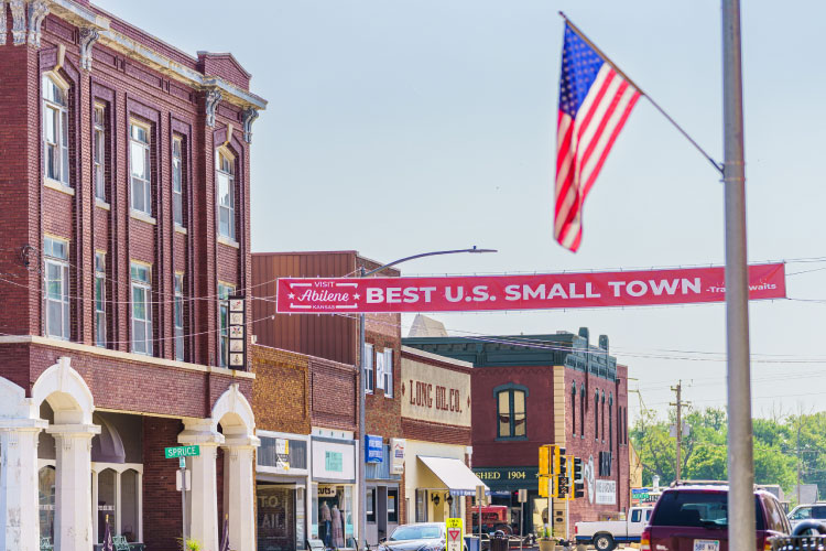 View of downtown Abilene, Kansas, on a sunny summer day. There’s a red sign stretched across the street, above the cars, that reads “Visit Abilene” and “Best U.S. Small Town.” 
