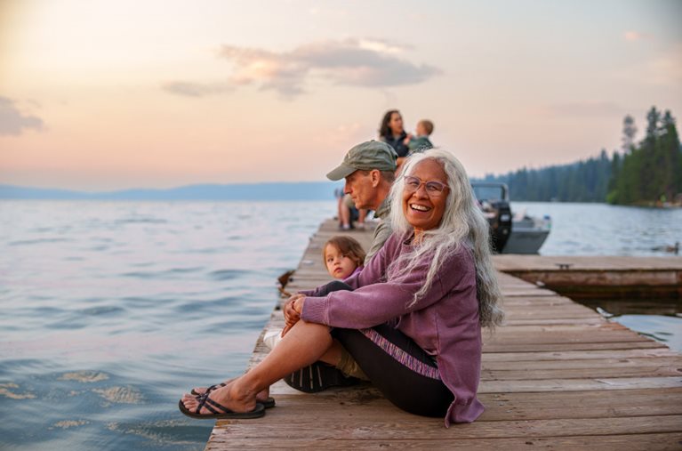 A retired woman is sitting on a dock with her husband and grandchild as the sun begins to set over the lake.