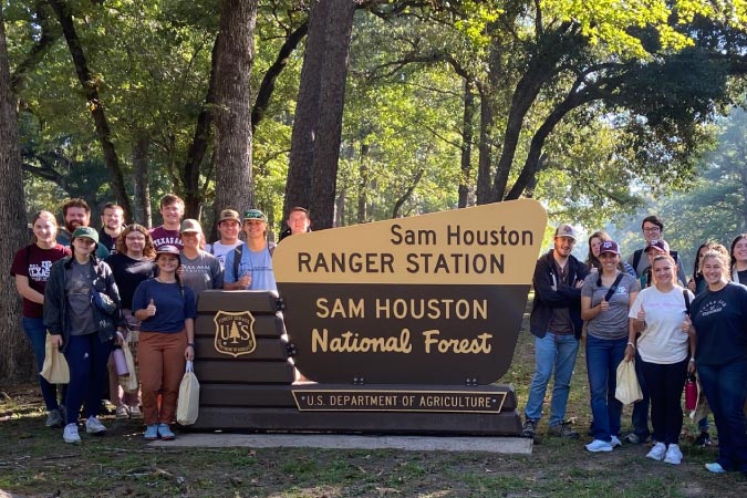 Students from Texas A&M University pose for a photo by a ranger station sign in the Sam Houston National Forest during a visit.