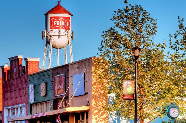 A view of the water tower in Frisco, Texas, behind a row of retro-looking storefronts in town.