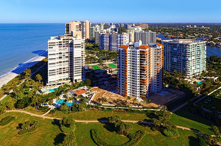 Tall condo buildings along Gulf Shore Boulevard beside the beach in Naples, Florida. Large grassy greenspaces dotted with tall palm trees surround the condo buildings