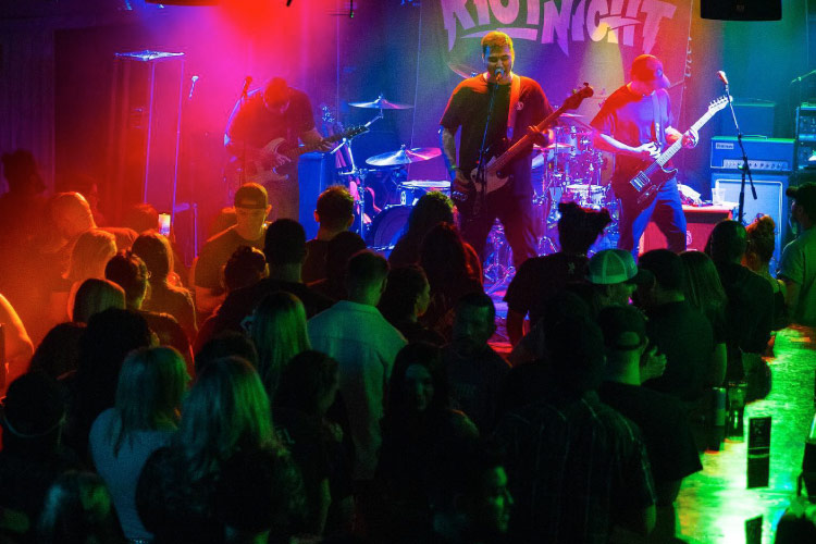 A live performance at the Rockbar Inc. music venue in Scottsdale, Arizona. Various stage lights cast orange, red, and green color on the stage as a rock band performs for a packed house.