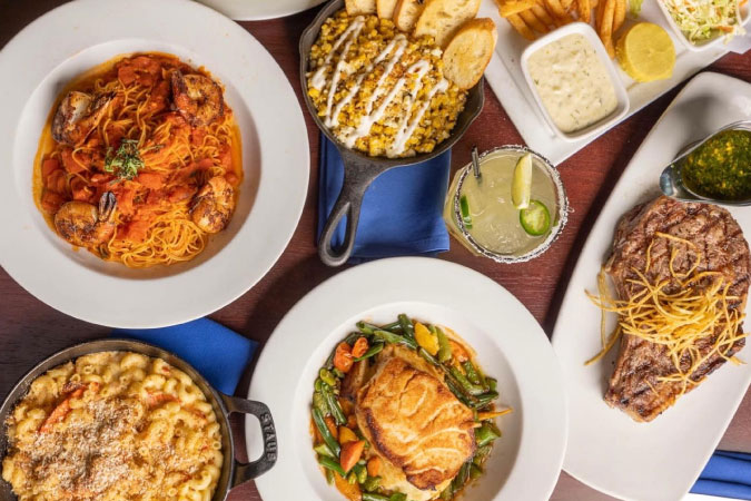View from above of a table full of entrees from Boathouse on the Bay in Long Beach, California, including steak, pasta, elotes, and more.