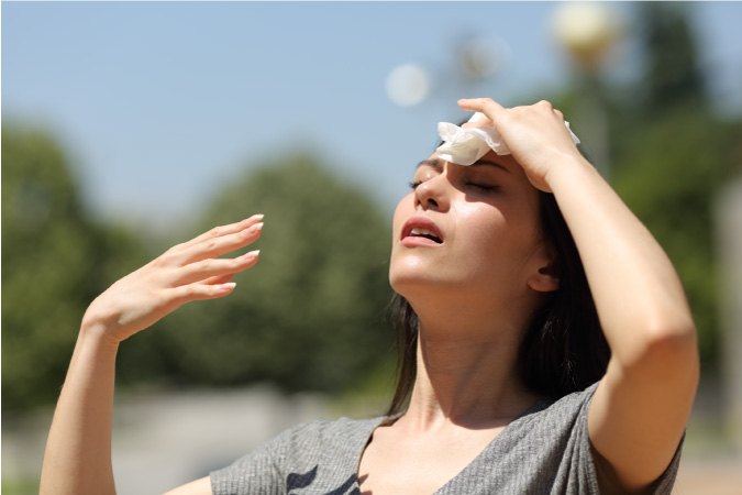 A woman is holding a cloth against her forehead as she fans her face with her hands on a hot, sunny day.