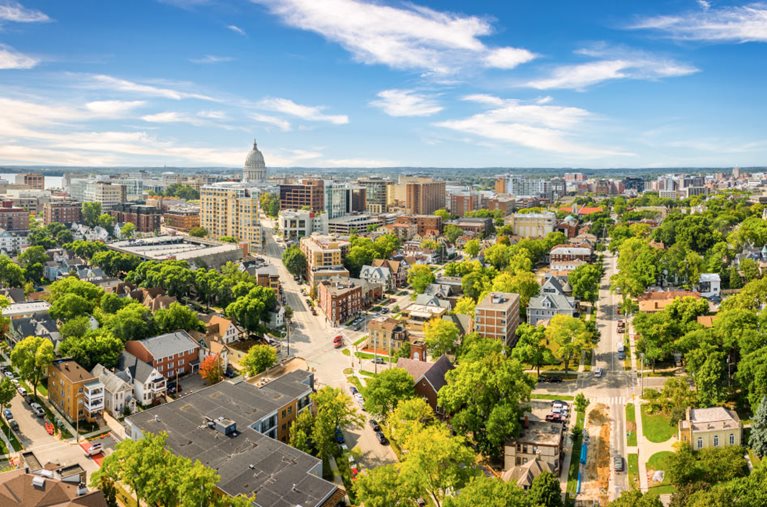 Aerial view of Madison, WI, on a sunny summer day. A diagonal city street runs toward a residential one, and lush green trees fill the gaps between buildings. In the distance, the Wisconsin State Capitol stands taller than any other building in the area.