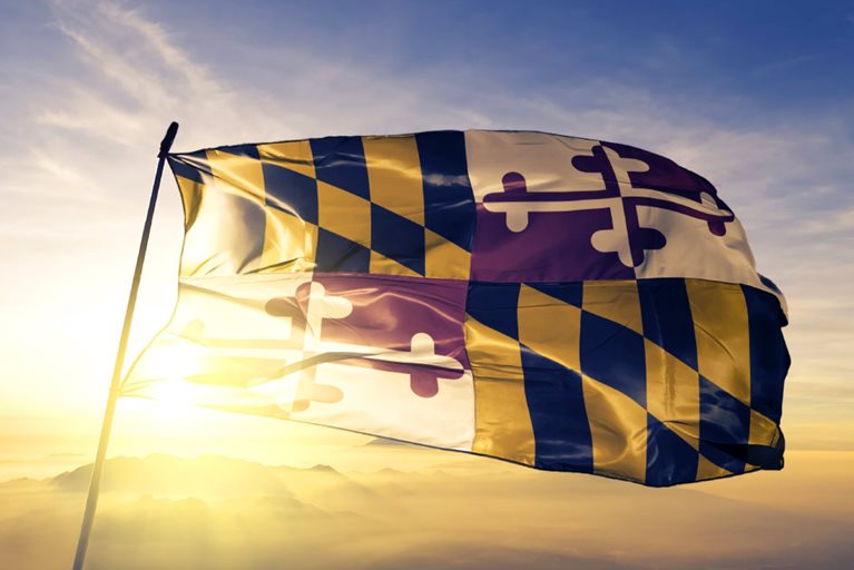 The Maryland state flag is blowing in the wind at the top of a flag pole while the sun is setting behind a range of mountains in the distant background.