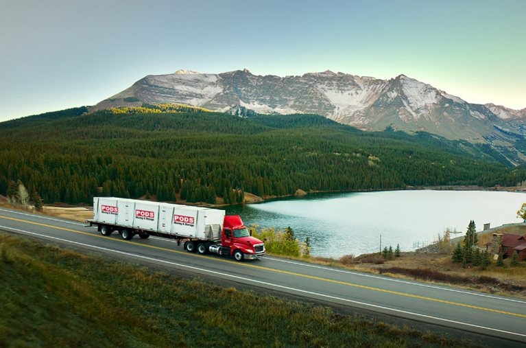 A PODS truck is transporting three PODS portable moving containers long-distance, passing a lake and mountain range.