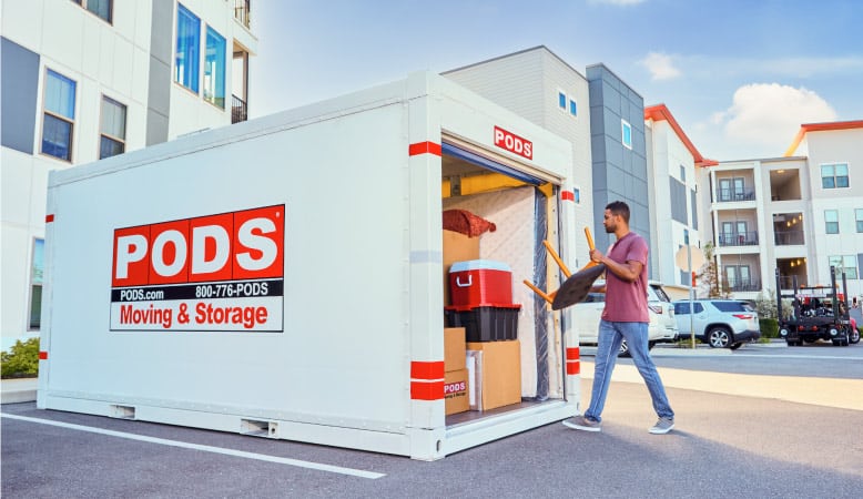 A man is loading a stool into his nearly full PODS portable moving container. It’s in a parking spot in an apartment complex. 