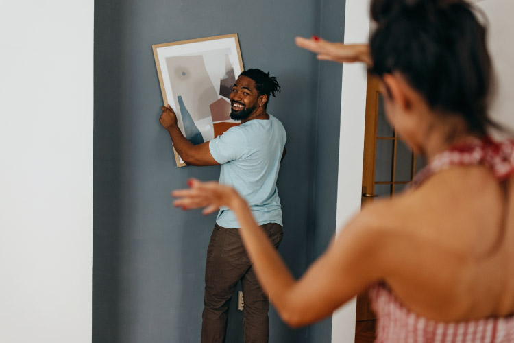 A man is holding a framed piece of abstract art on the wall. The art is tilted to the left, and his girlfriend is gesturing with her hands to show him how to turn it to make it level.