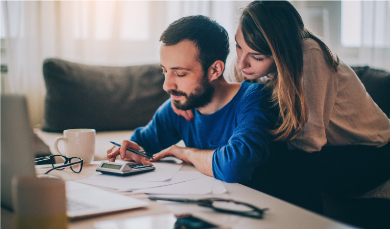 A man and woman are going over their budget together. The man is sitting on the floor at the coffee table, typing numbers into a calculator, and the woman is leaning over his shoulder.