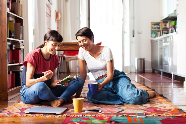 Two women who are roommates are sitting on the floor in their shared apartment, noting down the house rules in a little notebook.