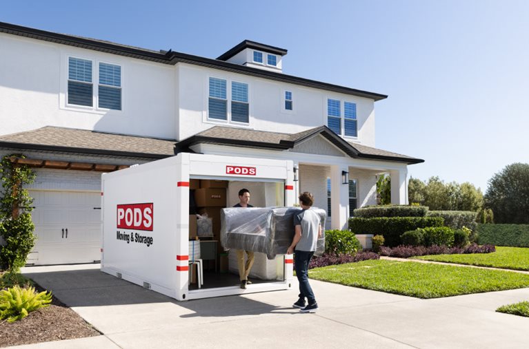 Two men are carrying an expertly wrapped couch into a PODS portable moving container in the driveway.