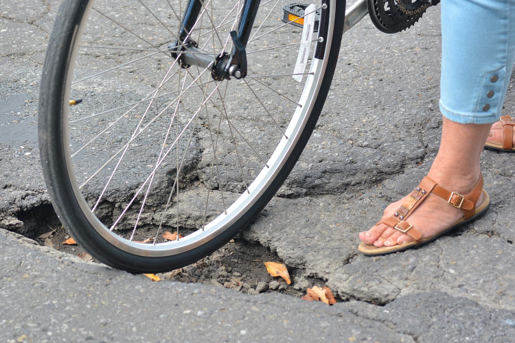 Close-up view of a woman’s sandaled feet and the front wheel of her bicycle as she navigates a large pothole in the road.Close-up view of a woman’s sandaled feet and the front wheel of her bicycle as she navigates a large pothole in the road.