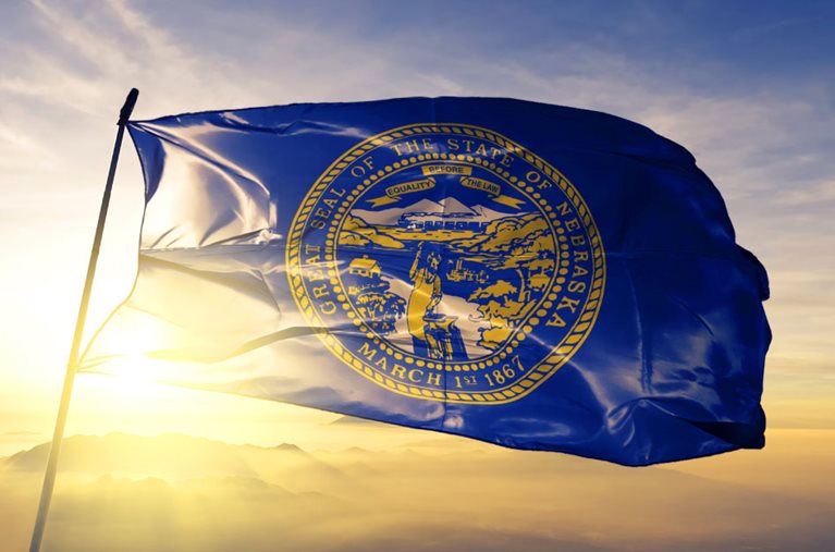 The Nebraska state flag is waving at the top of a simple flag pole as the sun rises behind a range of mountains in the distance.