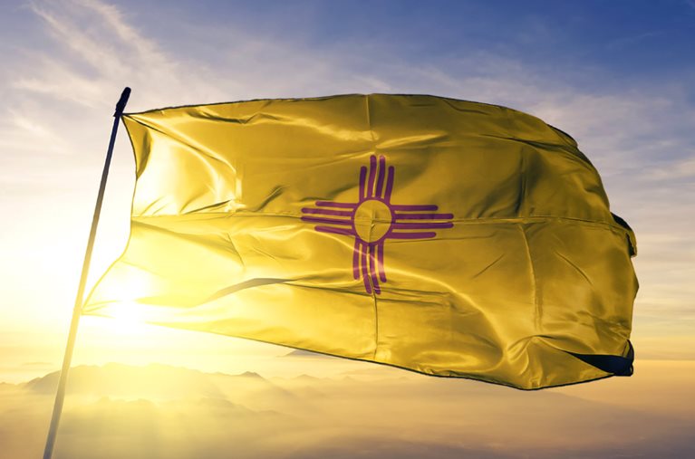 The state flag of New Mexico flies in the wind at the top of a flag pole as the sun rises behind a range of mountains in the distance. The flag features a yellow field with a red symbol in the center, representing the Sun.