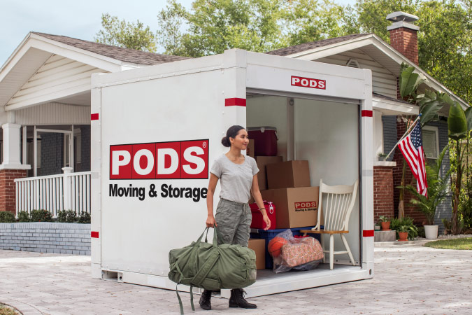 A soldier is holding her standard-issue green duffel as she stands beside a loaded PODS portable moving container, ready for her PCS move.