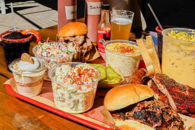 A delicious-looking BBQ spread from a restaurant in Dallas, Texas, featuring a brisket sandwich; ribs; sausages; various salads and slaws; and banana pudding