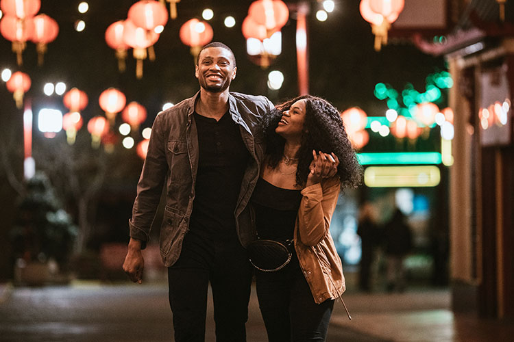 A smiling couple walk hand and hand through the streets of Los Angeles. Behind them are red lit Chinese lanterns that are out of focus.