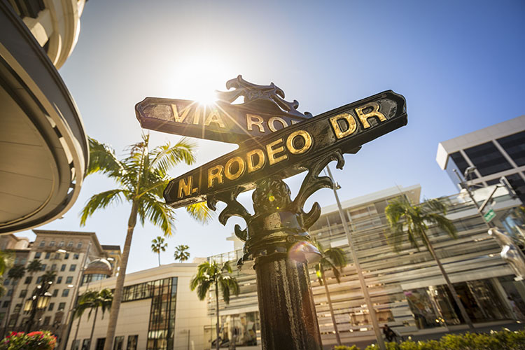 A sunny day in Los Angeles, California. The black street signs at the intersection of “Via Rodeo” and “N. Rodeo Drive” are in the forefront, with high-end stores in the background.