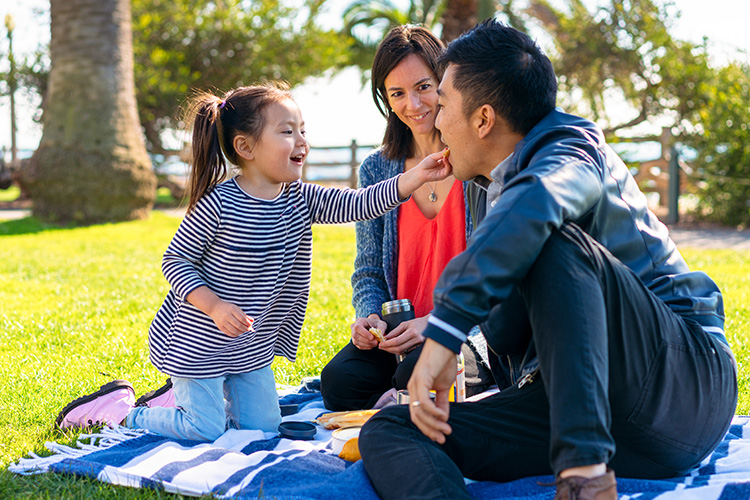 A young family (mom, dad, and toddler) sit on a blue and white striped blanket in a park in Los Angeles. The young girl is feeding her dad a snack. 