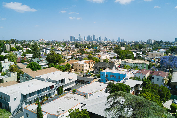 A bird’s-eye view of a neighborhood in Los Angeles. The buildings have flat roofs that are different colors. In the background is the skyline of Los Angeles. 