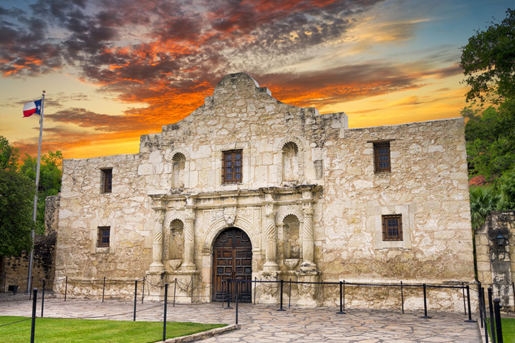  Image of the Alamo in San Antonio at sunset. The sky is yellow and orange. The Alamo is white stone with a flagpole with the Texas flag off to the side of the building. 
