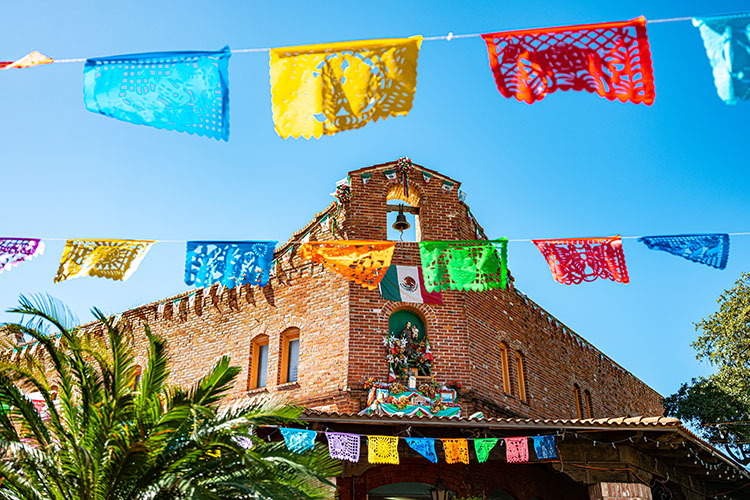 An image of a market in San Antonio, TX. There is a red-and-brown brick building adorned with multi-colored flags and flowers. In the foreground are multi-colored flags hanging in two rows. 