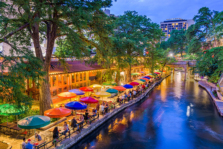 An image of the River Walk in San Antonio. It is dusk and to the left are tables and chairs under different colored umbrellas. To the right is a stone walking bridge.