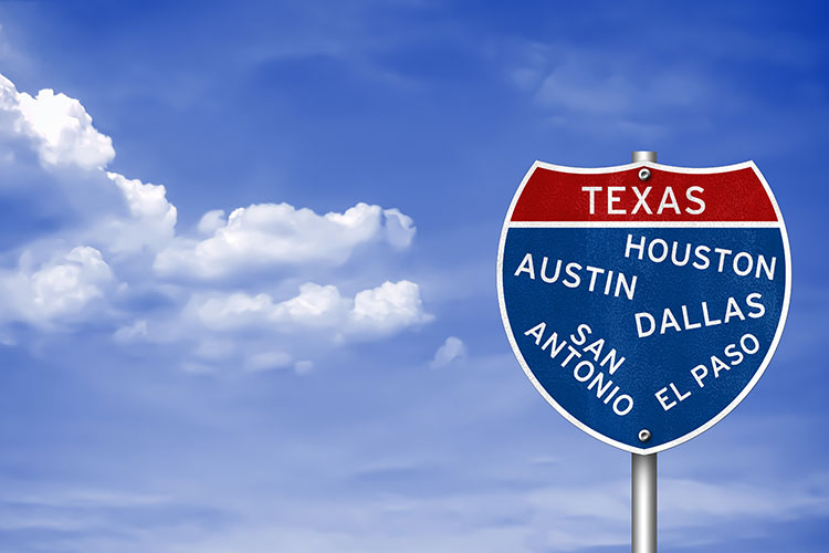 A clear sky with white clouds. To the right is a sign that reads “TEXAS” in white text in a red box. Underneath is a Blue box that reads “HOUSTON,” “AUSTIN,” DALLAS,” SAN ANTONIO,” and “EL PASO” in slanted text. 