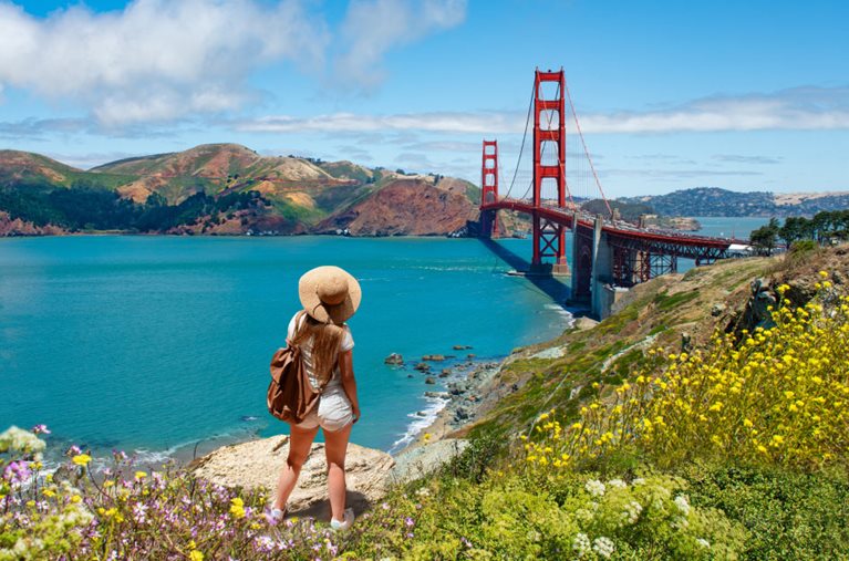  A young woman in a sun hat is standing on a cliff above San Francisco Bay, looking out over the Golden Gate Bridge on a sunny day.