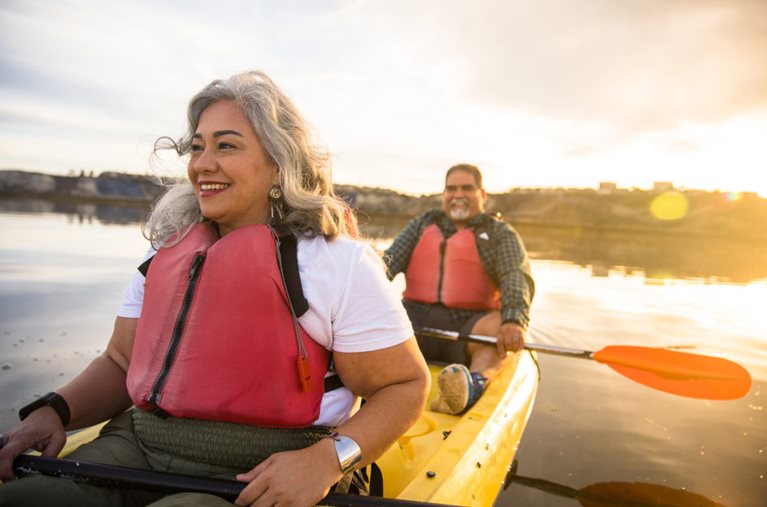 A happy, mature couple enjoys kayaking on a lake during golden hour. The kayak is yellow, and they’re both wearing red life jackets.