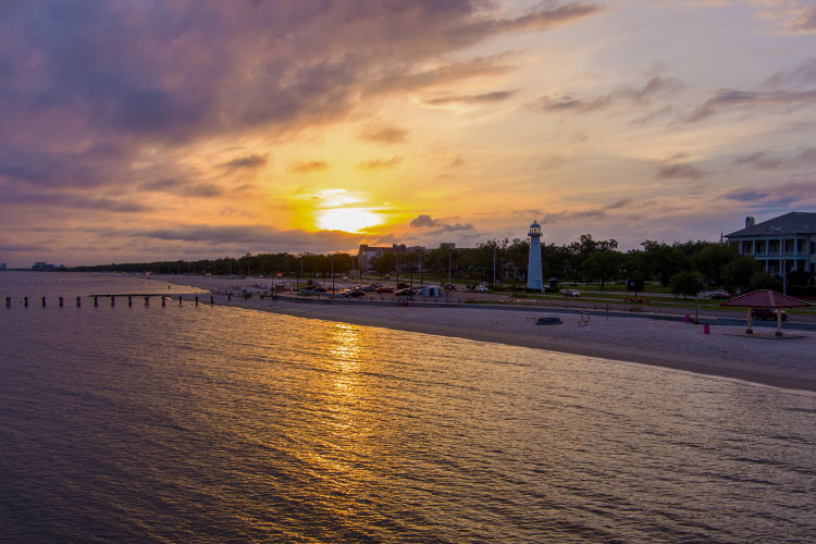 View of the Gulf Coast waterfront in Biloxi, Mississippi, at sunset. The sun sets in the distance, casting yellow light across the coastal waters.