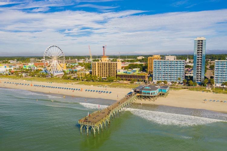 Aerial view of the beach in Myrtle Beach, South Carolina, on a summer day. The beach is lined with tall condos and hotels, and there’s a Ferris wheel to the side. Blue umbrellas dot the beach, and visitors explore the long pier that juts out into the surf.