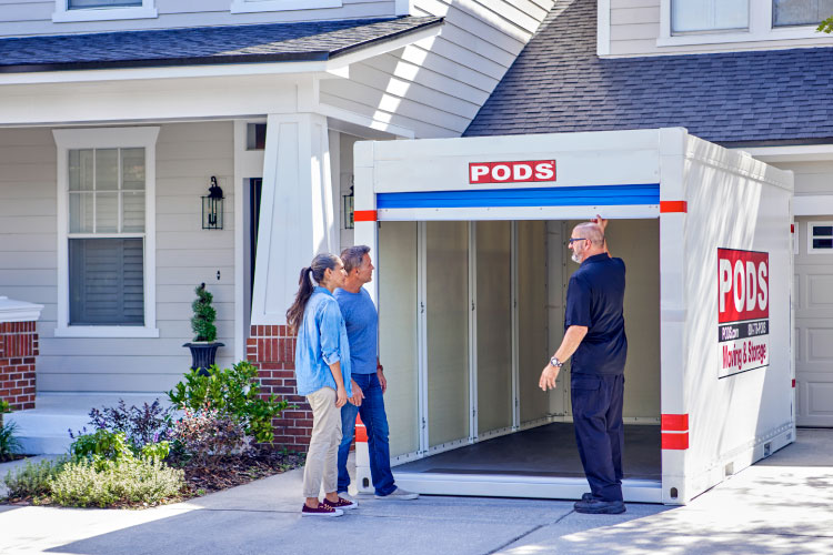 A PODS driver is standing in a residential driveway, opening the door of a PODS portable moving and storage container so a mature couple can see inside.