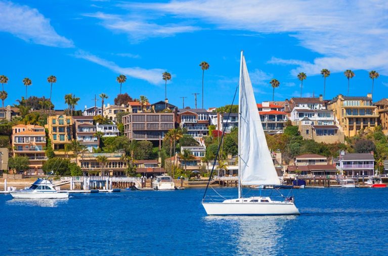 A sunny summer day at Newport Bay in Newport Beach, California. The waterfront is packed with condos and commercial buildings, as a small sailboat glides past the coast.