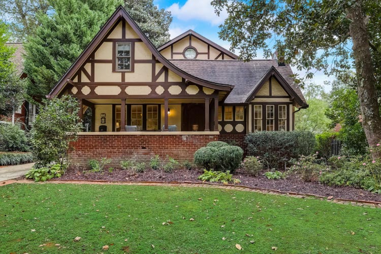 A historic Tudor home in Avondale Estates, Georgia, is nestled between mature trees and features a lovely covered porch.