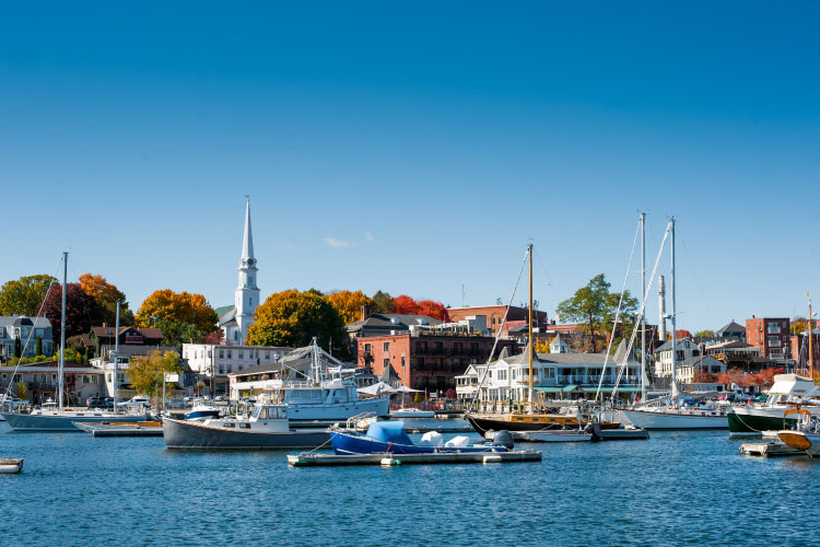 Several boats are moored in the harbor in Camden, Maine. Beyond the harbor, the city can be seen beneath a clear blue sky.