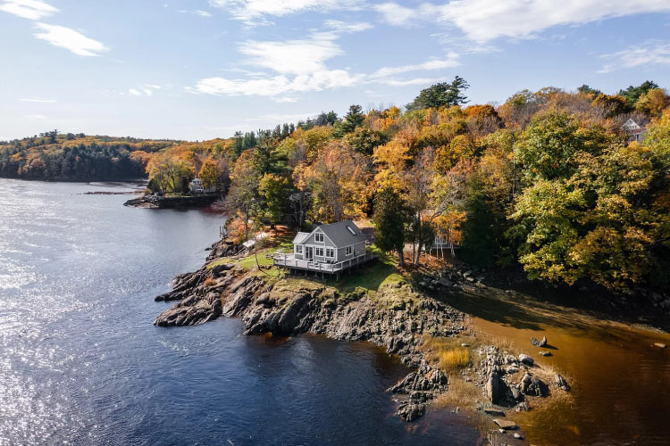 A two-story home with a large porch on the waterfront in Hampden, Maine. The home sits atop a rocky outcrop, jutting out from a heavily wooded area. The water surrounding the property is dark and placid. 